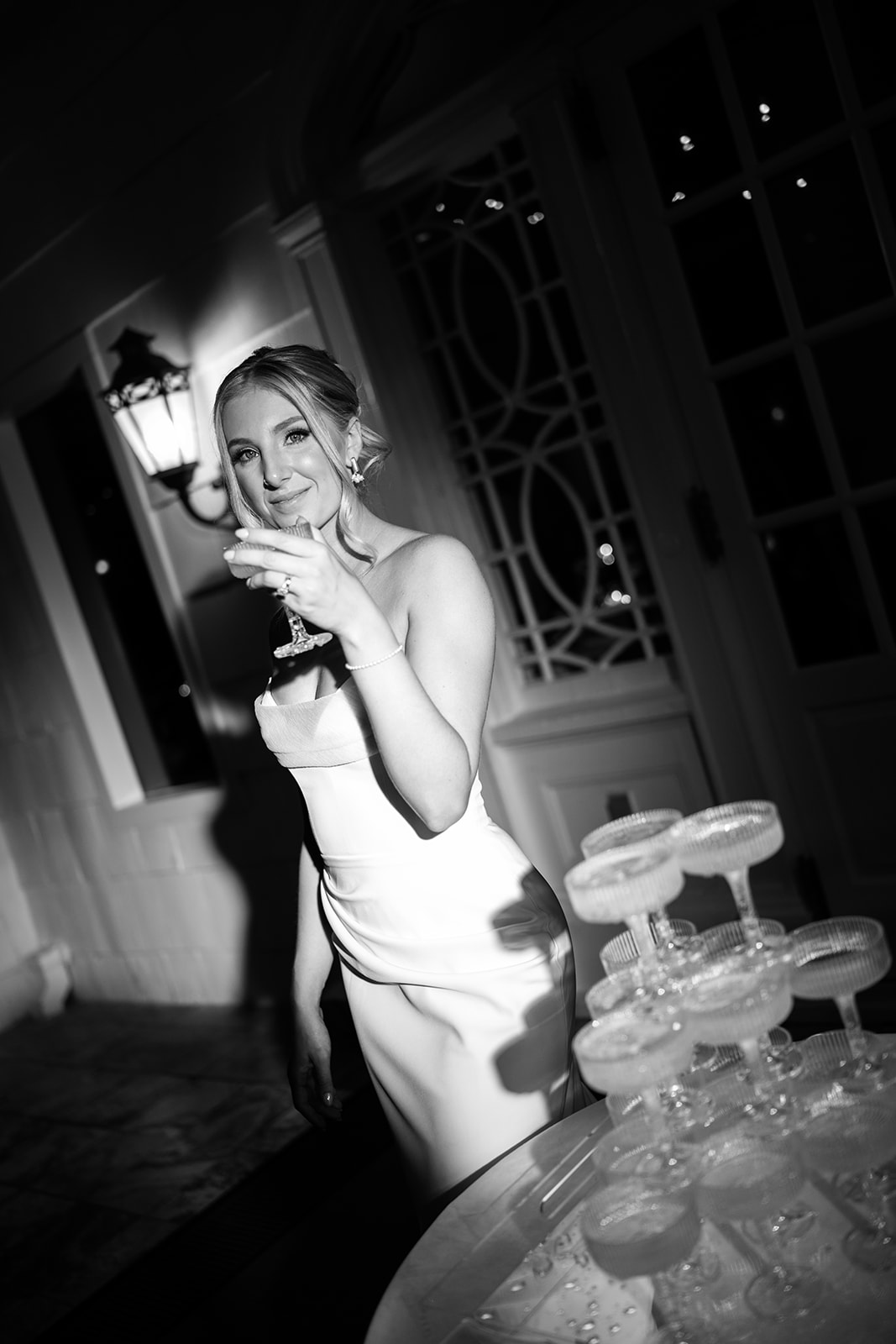 Black and white portrait of the bride raising a champagne coupe glass and smiling at the camera.